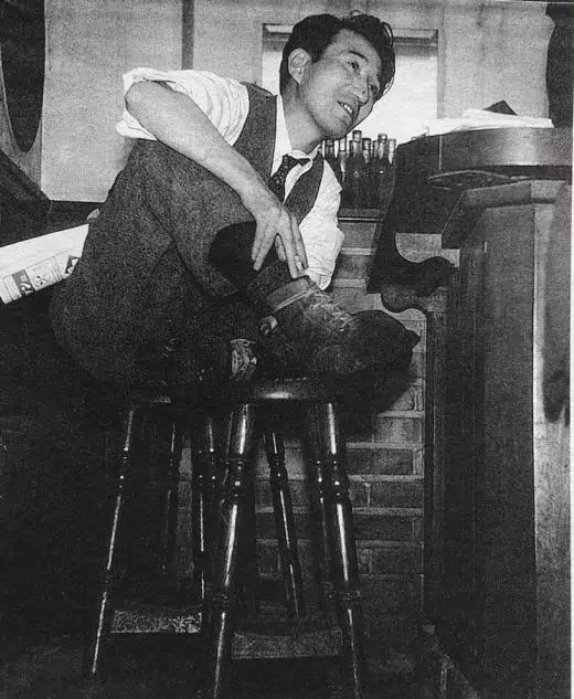 Photographie en noir et blanc de DAZAI assis en tailleur sur un tabouret de bar.